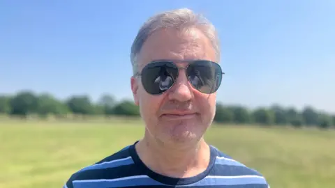 BBC / Elizabeth Baines A man with grey hair smiles at the camera. He wears sunglasses and a blue striped top. Behind him green space is blurred.