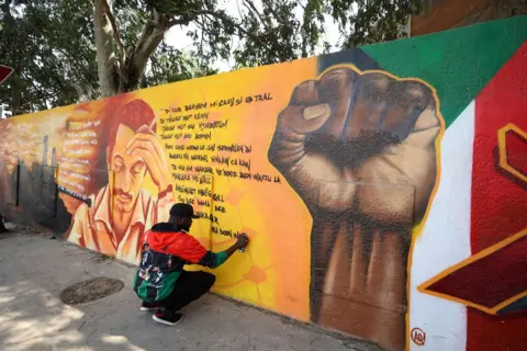 CEM OZDEL / GETTY IMAGES A man paints graffiti text in the Wolof language onto a large mural. Next to it is a clenched fist, symbolising defiance.