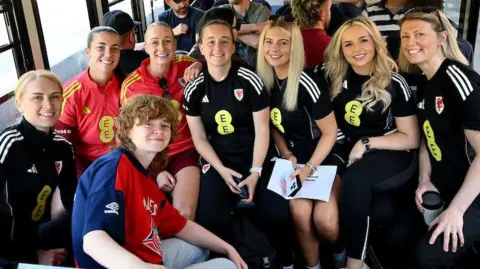 A group of women dressed in black and red football kits, all looking at the camera and smiling