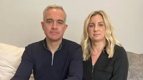Ralph with short white hair, wearing a dark zip-up top and Lauren, with long blond hair also wearing a dark zip-up top. They are looking at the camera and are sitting on a light-coloured bed or sofa.