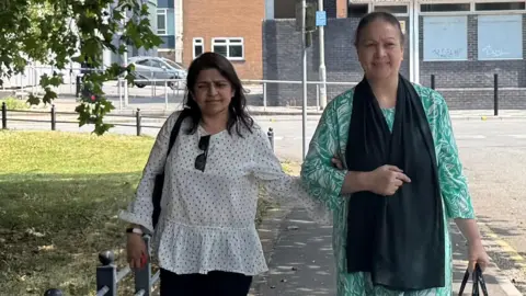 Two women are pictured walking down a pavement towards the camera, the woman on the left is wearing a white blouse and black trousers. The woman on the right is wearing a green dress. It is a sunny day.