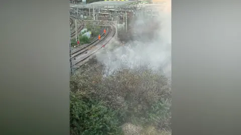 London Fire Brigade Smoke pours from bushes close to a railway line in an urban area. 