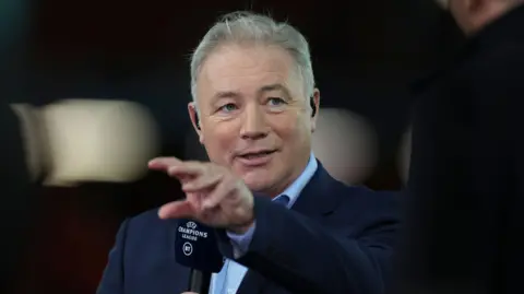 Ally McCoist is holding a microphone and talking into it at a sport event. He is moving his hands in front of his face while he is talking.