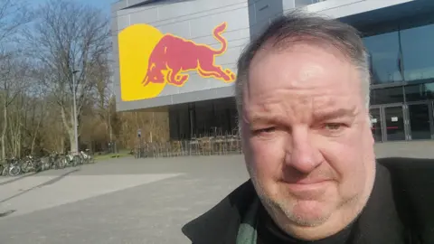 James Parsons, a man with short hair and stubble, is pictured in front of a football ground with a large red bull insignia - the symbol for energy drink Red Bull. 