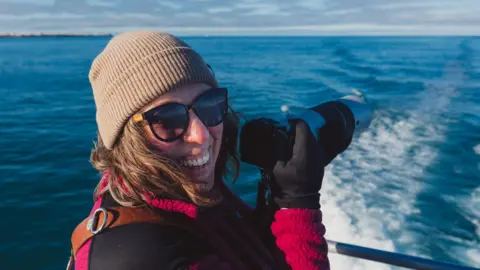 Rebecca Douglas wearing a beanie and fleece, smiling at the camera. She is on a boat and pointing a camera out to sea.