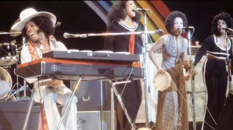 Getty Images Sly and other members of Sly and the Family stone sing on stage in front of a rainbow backdrop.