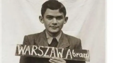 Liron Velleman A black and white photo from 1945 showing Alec Ward holding up a chalk board bearing his birth name Abram Warszaw. He is wearing a collar shirt and a jacket and he has dark parted hair.