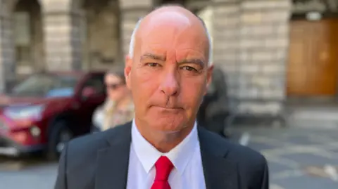 Tommy Sheridan standing outside the Court of Session - he is dressed in a black jacket, white shirt and red tie. He is mostly bald with white hair.