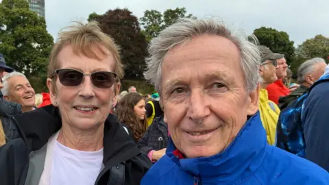A older man and woman, it is a head and shoulders shot of them. She has short, light-brown hair and has dark shades on. She has a dark coat. The man on her right also has short grey hair and a blue coat zipped up. Both are looking at the camera.