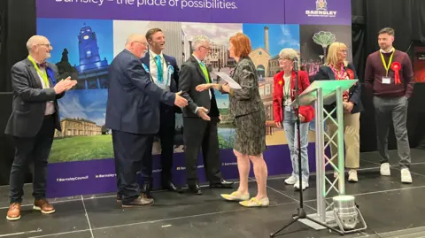 BBC/Victoria Scheer A group of people standing on a raised indoor stage in front of a large purple display board. The display reads “Barnsley – the place of possibilities” and includes the Barnsley Metropolitan Borough Council logo, along with a collage of photographs depicting buildings, landmarks, and public spaces.
