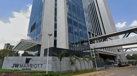 Google is a gray outer with a modern hotel, with a lot of floors to roof windows. Outside, a sign of JW Marriott Nairobi says. There is some greenery in front of it. 