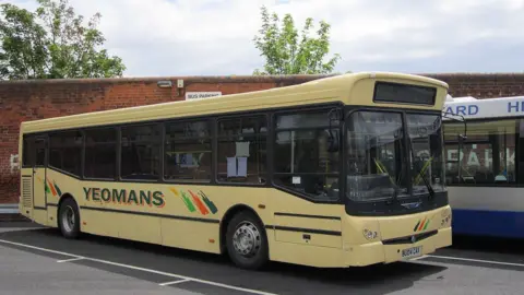 A pale yellow single decker bus is parked in a car park. On the side is has "Yeomans" in bold dark green and orange font.