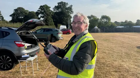 An older man with short, grey hair and wearing a yellow hi-vis jacket is standing in a field. The grass is dry and he is holding a drone controller. He is smiling and standing side on with his head turned towards the camera. There are a few cars parked in front of where he is standing.