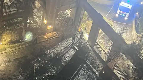 The picture shows rubble and damage following an explosion outside the hotel. The wreckage is blackened in the foreground and an emergency vehicle with its blue lights flashing can be seen in the background.