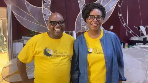 Lily-May Symonds/BBC A man and a woman stand together to pose for a picture. The man is bald and wears black rimmed glasses and a yellow T-shirt with the words, UK Centre for Carnival Arts on it. He rests one of his hands on his hip. The woman has short black hair and also wears black rimmed glasses along with the same yellow T-shirt with a denim cardigan over the top.