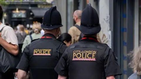 The backs of two people wearing black police uniform that reads City of London Police, including tall hats. They are walking on a busy street.