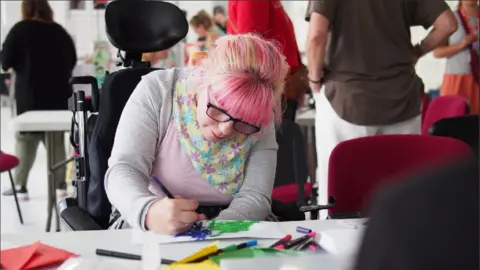 A woman with pink hair and glasses and in a wheelchair leans across a piece of paper on which she is colouring with a blue felt tips, with other felt tips on the table 