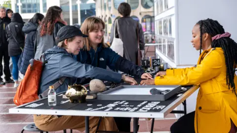 Kate Green Photography A group of people, including a woman in a yellow, and two other people, with their arms outstretched lacing white letters on a board