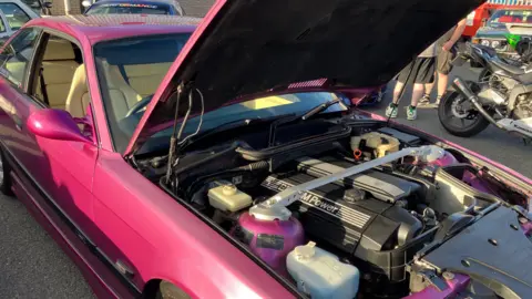 A hot pink modified car parked with the bonnet open displaying the modified engine.