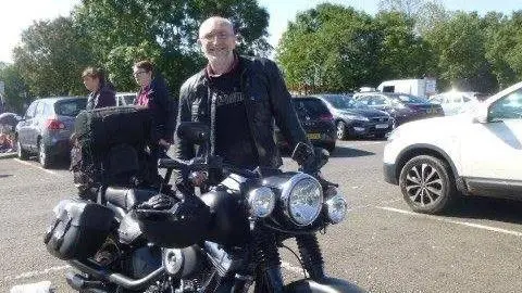 Spindrift A man with a bald head and glasses in a leather jacket stands next to a large motorbike