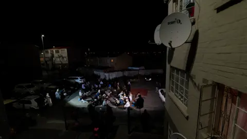 BBC/Shiraaz Mohamed A woman looking down from a flat window at people gathered for dhikr in Manenberg, Cape Town - South Africa