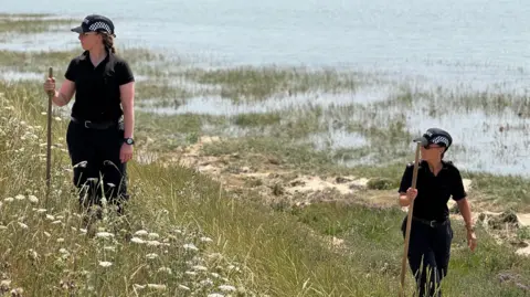 Zoie O'Brien/BBC Two female police officers conducting a search on the seafront amongst some grass and a beach.