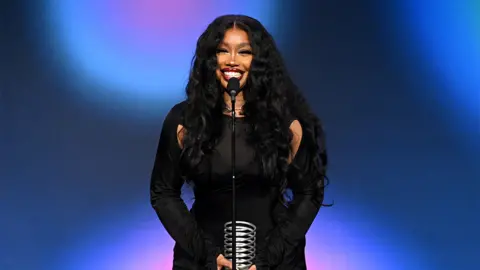 Getty Images SZA, a woman wearing a black outfit, standing on stage behind a microphone, accepting an award. She is smiling. The background is dark and light blue and pink.