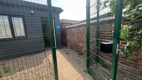 Green metal gate with grey wood panelled building cabin behind it