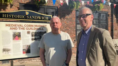 Two men stood in front of an information board. The information board reads: "HISTORIC SANDWICH / MEDIEVAL CORN MARKET AND THE GUILDHALL".