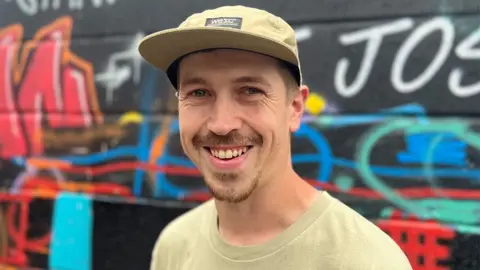 Head and shoulders shot of Shawn Sharpe standing in front of a graffiti wall. He is wearing a beige cap and t-shirt, has a short dark goatee and is smiling