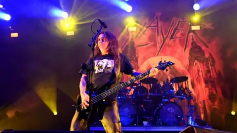 Singer Tom Araya of the band Slayer performs onstage during the bands final show of the "Final Campaign" tour. He is dressed in all black and has long, wavy brown hair and is playing the guitar. A drum set can be seen behind him in front of the band's logo