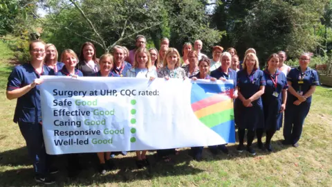 University Hospitals Plymouth NHS Trust Staff from Derriford Hospital stand on a lawn in front of trees and hold up a banner saying CQC has rated surgery services at the site as good. The word good is written five times in green along with five green dots. Many of the staff are wearing scrubs.
