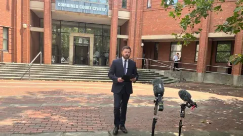 A man in a suit stands outside Hull Crown Court, a large red brick building, with microphones in the foreground.