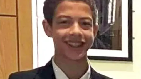 Noah Donohoe, wearing a white shirt, black and green tie, and a black suit jacket. He has short brown hair and is smiling at the camera. 