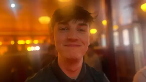 A young man with short brown hair smiles to the camera. He wears a green polo buttoned up fully. Behind him, out of focus, is a bar where a table of people are drinking.