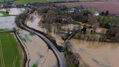 Eddie Mitchell A curving country road through fields and a small collection of homes, seen from above. The fields and part of the road are flooded with brown water.