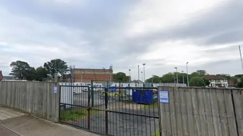 A tall wooden fence with an iron gate in the middle. Through the gate, caravans and camper vans are parked on a forecourt and behind that there are floodlights and a brick building.