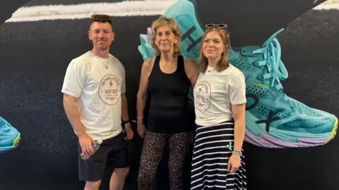 David Edwards A man and two women stand together in a line and smile at the camera. The man wears a white T-shirt and black top. He is holding a water bottle. He has sunglasses resting on top of his head. A woman in the middle has short blonde hair and is wearing a black gym tank top and animal print leggings. The woman on the right has short brown hair, sunglasses on her head, is wearing a white T-shirt and a striped black and white long skirt.