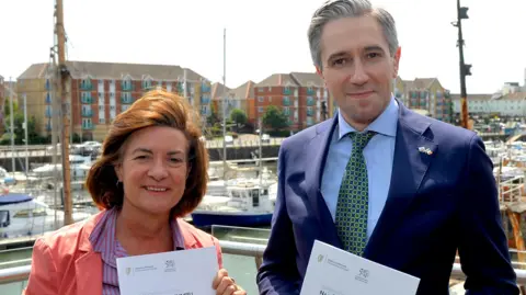 Welsh Government A brown haired woman with a red suit jacket and a stripy blue and red shirt holding an A4 document alongside a man wearing a blue suit, blue shirt and tie holding an A4 document with boats in a harbour in the background