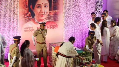 Reuters Actor Tabu wearing a white kurta and a scarf pays her respects to Indian singer Asha Bhosle - who is seen lying in a casket wrapped in the Indian flag, as police officers and family members stand nearby