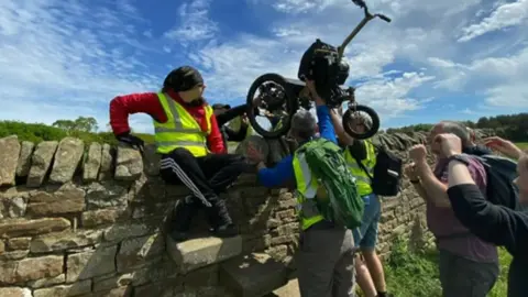 Brynn Hauxwell Brynn's wheelchair is lifted over the wall by his support team