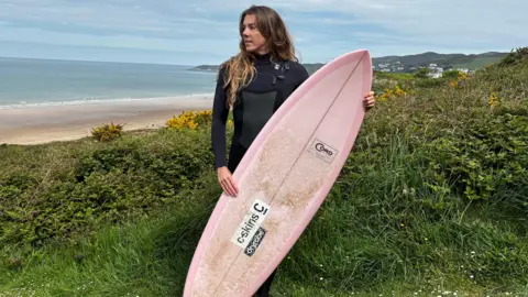 A person in a wetsuit is standing on grassy cliffs above a beach, holding a pink surfboard. In the background, there is a wide sandy bay, calm sea, and distant hills under a cloudy sky, with yellow flowers and greenery in the foreground.
