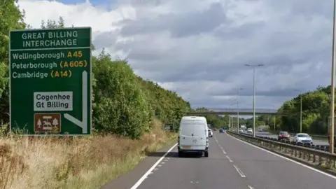 Google Dual carriageway road with a green sign to the left direction traffic to a turn to Cogenhoe and Great Billing. The heading says GREAT BILLING ROUNDABOUT. A white van is heading away from the camera on the eastbound carriageway while a lorry and cars are visible on the westbound.