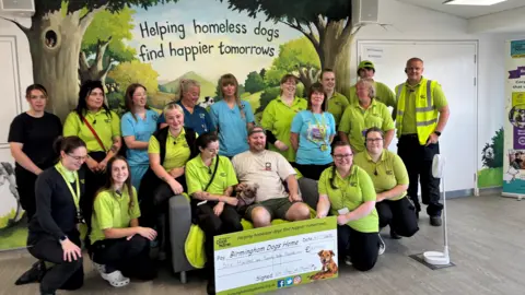 Birmingham Dogs Home Peggy – a pug and Chinese crested cross dog - sits on a sofa with her owner and is surrounded by a group of 17 volunteers from Birmingham Dogs Home, most of whom wear light green charity shirts. They are sitting and standing in front of a wall decoration reading "Helping homeless dogs find happier tomorrows"