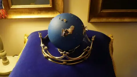 Qays Najm/BBC The navy-coloured globe is set in a golden metal crown. The item sits on a purple velvet cushion. The room is lit by a lamp and pictures in gold frames are on the wall behind it. 