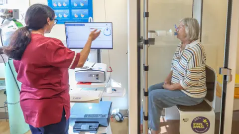 Patient Kirsty Dudin sits in a booth and breathes into a lung function testing machine. She is sitting on a plastic seat which says "Stars Appeal". Outside the booth, a doctor monitors the feedback from a computer.