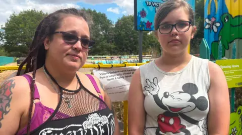 A mother and daughter, both wearing glasses stand in front of a locked gate to the Victoria Embankment Paddling Pool. There is a colourful temporary storage building behind the girl and the drained paddling pool beyond.
