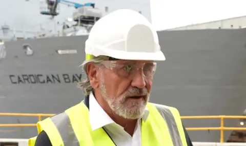 A man with a short white beard wears a white hard hat, clear safety glasses and a high‑visibility vest. He stands in front of a large docked ship with “Cardigan Bay” painted on its side.