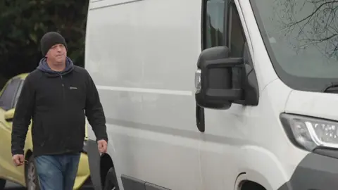 Dave wearing a black beanie and coat walks up to the door of his white van.
