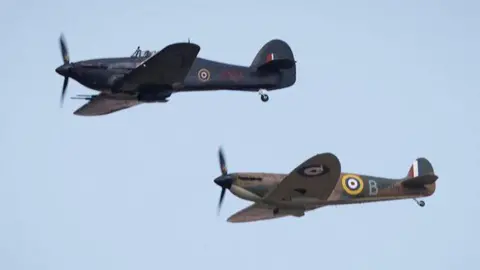 MOD Two spitfires fly, one above the other in the blue sky. The famous red, white and blue roundel emblem can been seen on the back of the plane.